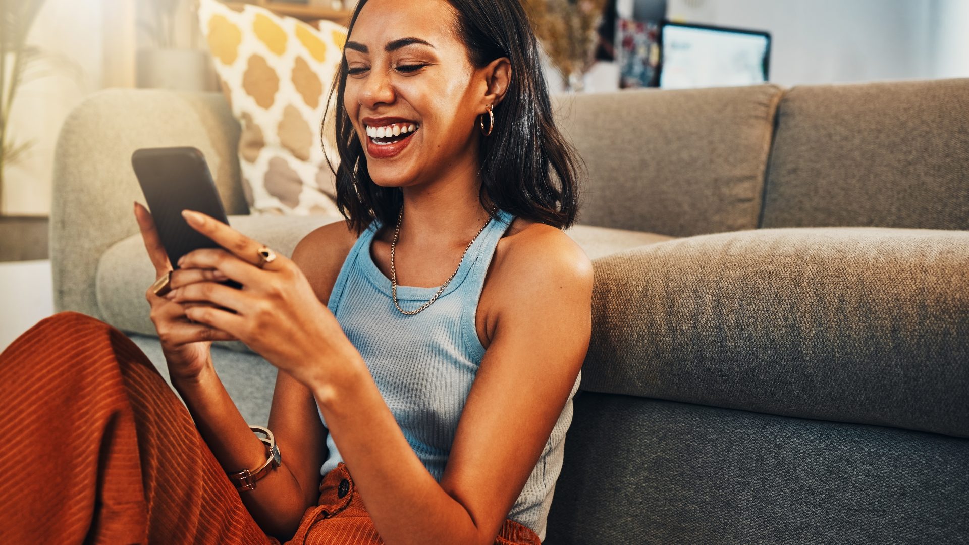 Beautiful mixed race woman browsing internet on cellphone in home living room. Happy hispanic sitting alone on floor in lounge and using technology to network. Laughing while scrolling on social media