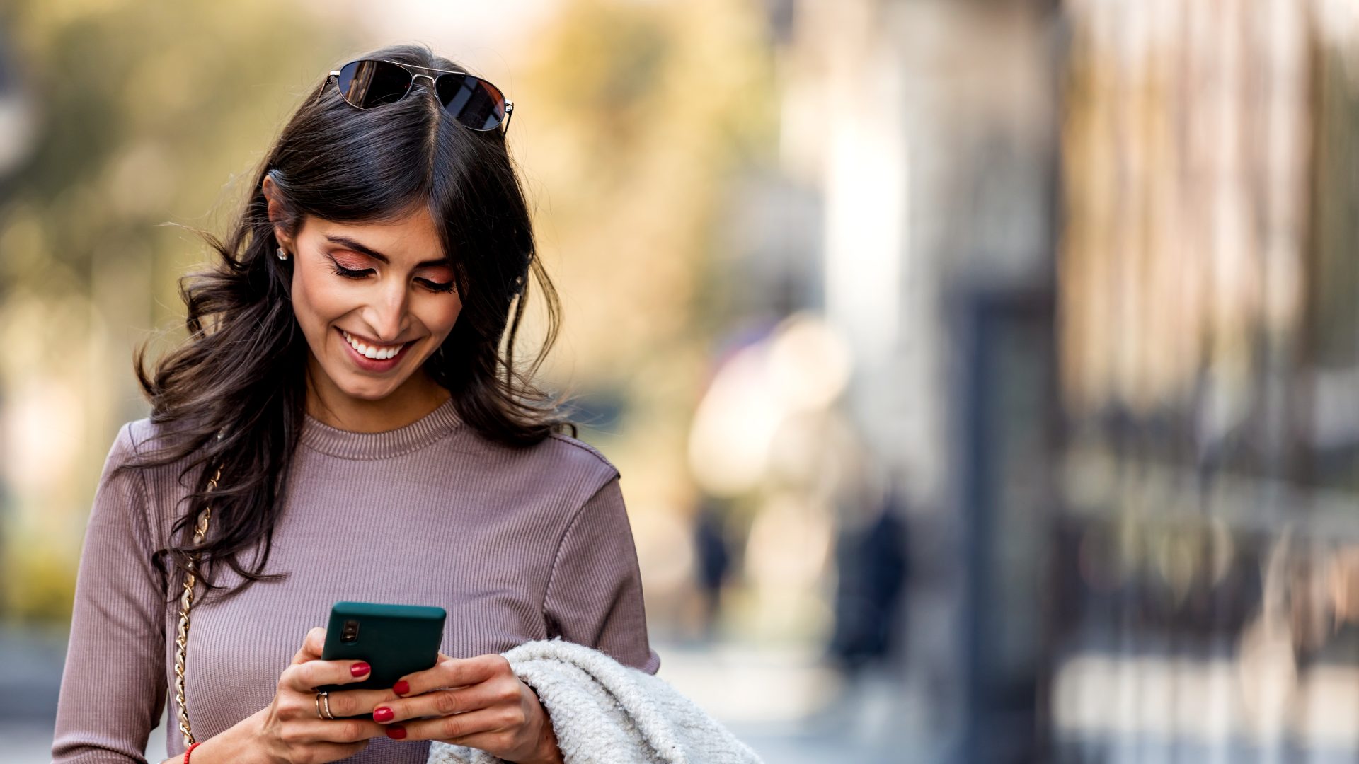 Beautiful mid adult woman walking and texting message on mobile phone outside business center.