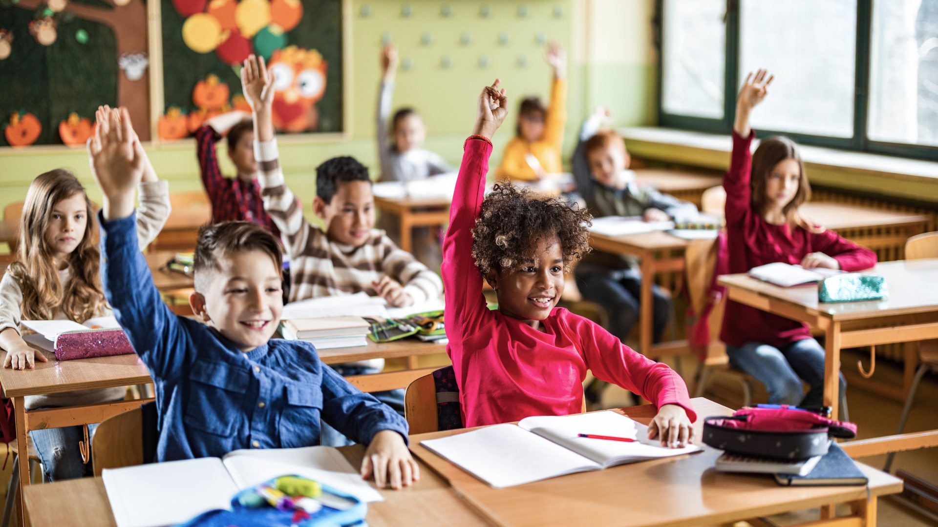 Happy elementary students raising their hands on a class at school.