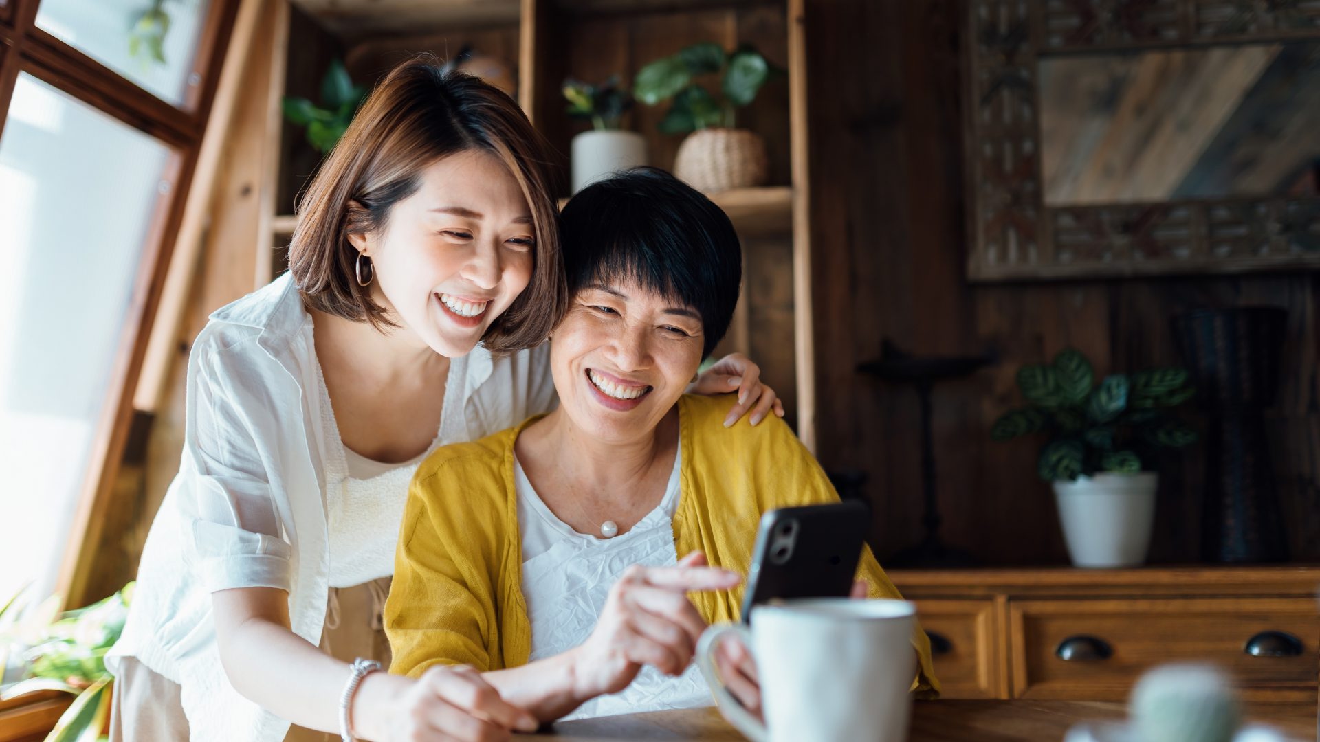 Affectionate Asian senior mother and daughter using smartphone together at home, smiling joyfully, enjoying mother and daughter bonding time. Multi-generation family and technology