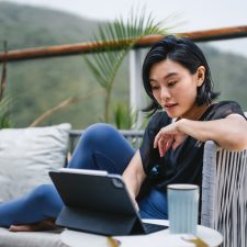 Young Asian woman sitting on the balcony, using digital tablet and having a cup of coffee in the morning. Young female using computer for leisure or business at home