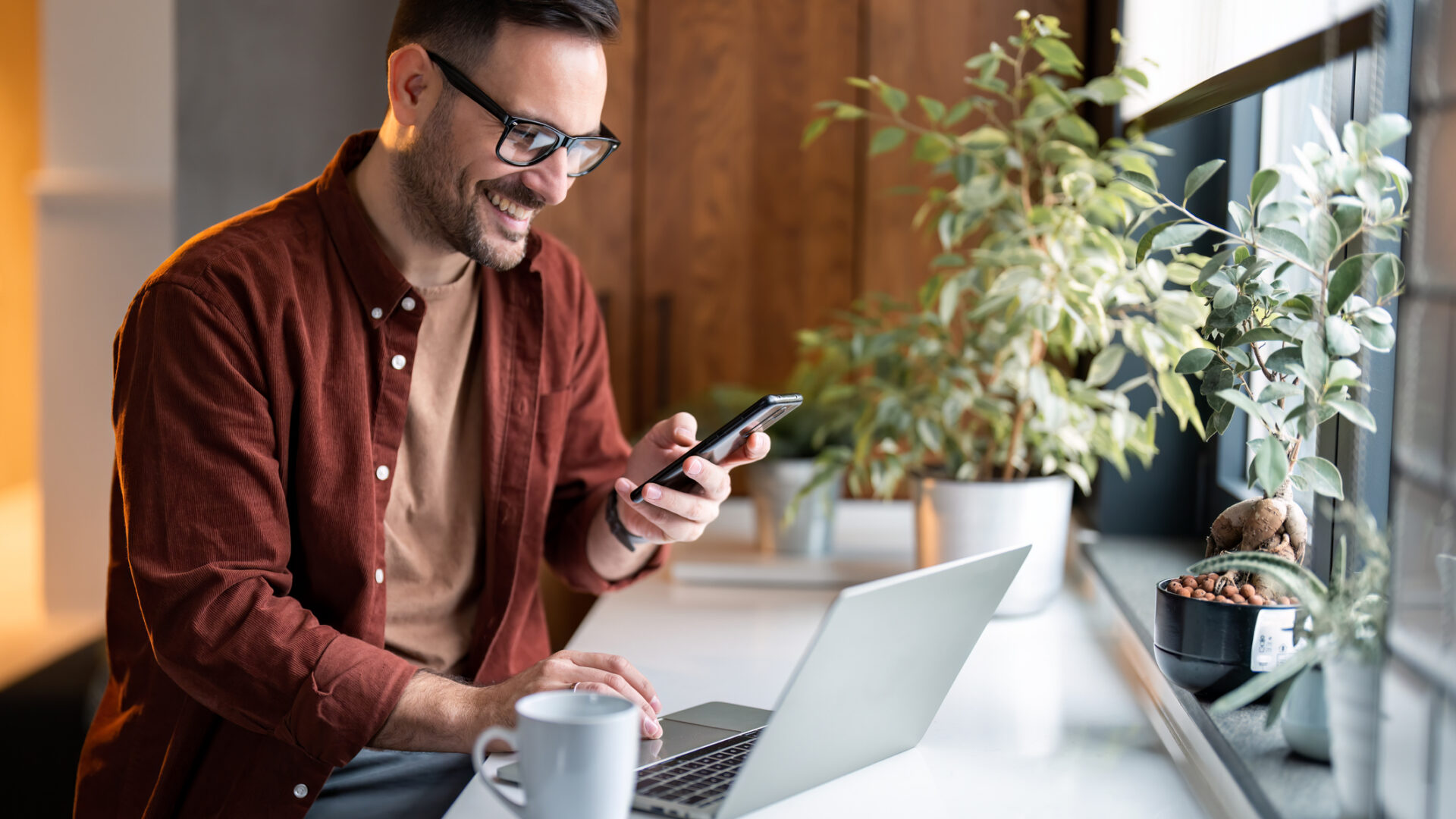 Satisfied modern millennial man in stylish casual clothes using smartphone and laptop computer for electronic banking, making reservation, online shopping and payments while spending time at home.