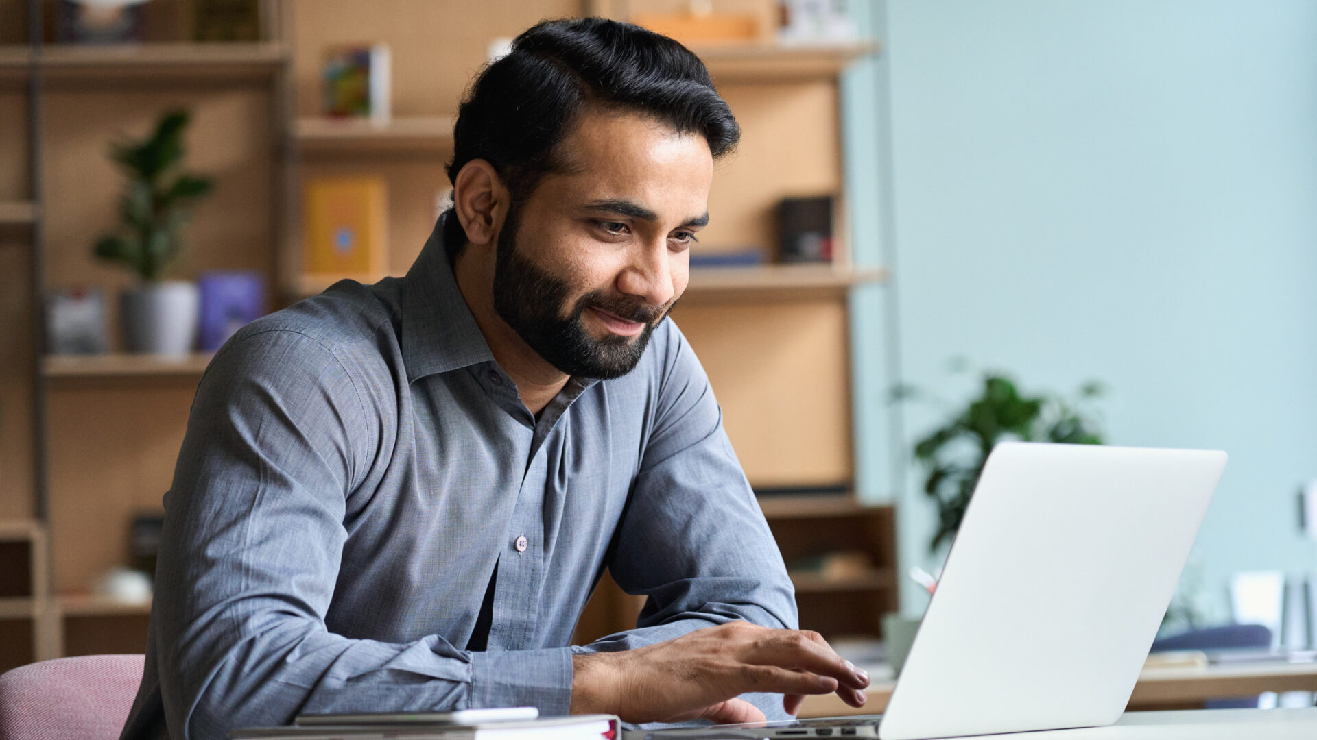 Smiling indian business man working on laptop at home office. Young indian student or remote teacher using computer remote studying, virtual training, watching online education webinar at home office.