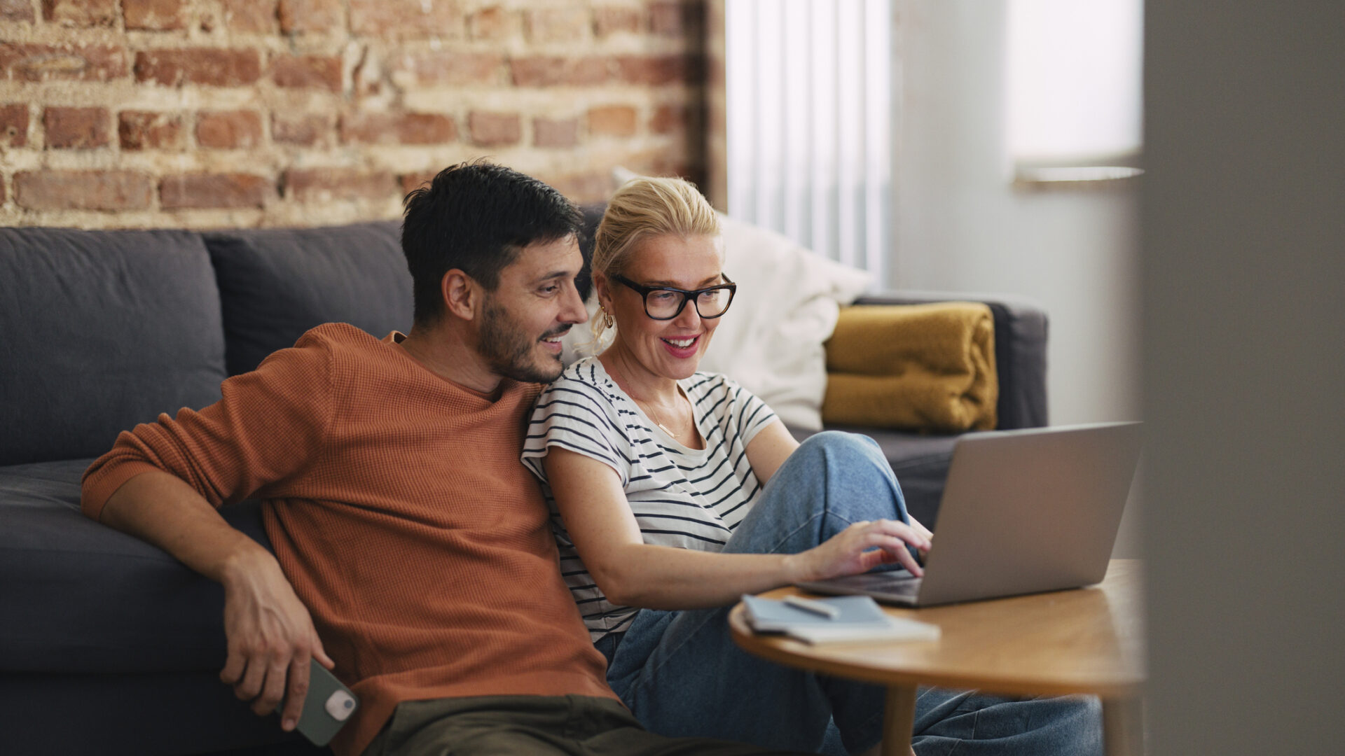 Happy couple sitting on the floor cuddled together watching a show or a movie on the laptop computer. They are smiling while looking down at the screen.