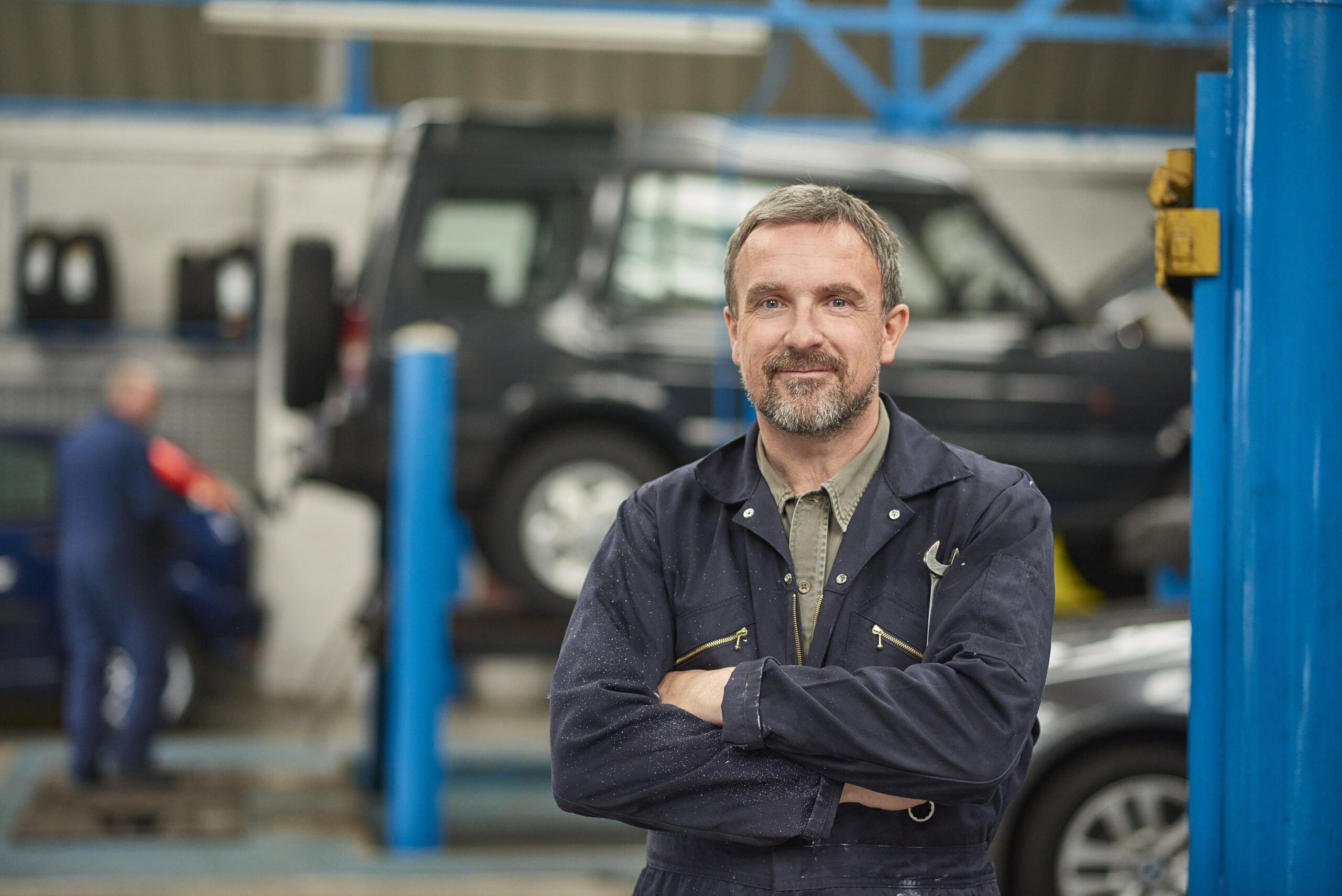 A smiling garage owner stands looking to camera from his busy auto repair shop .
