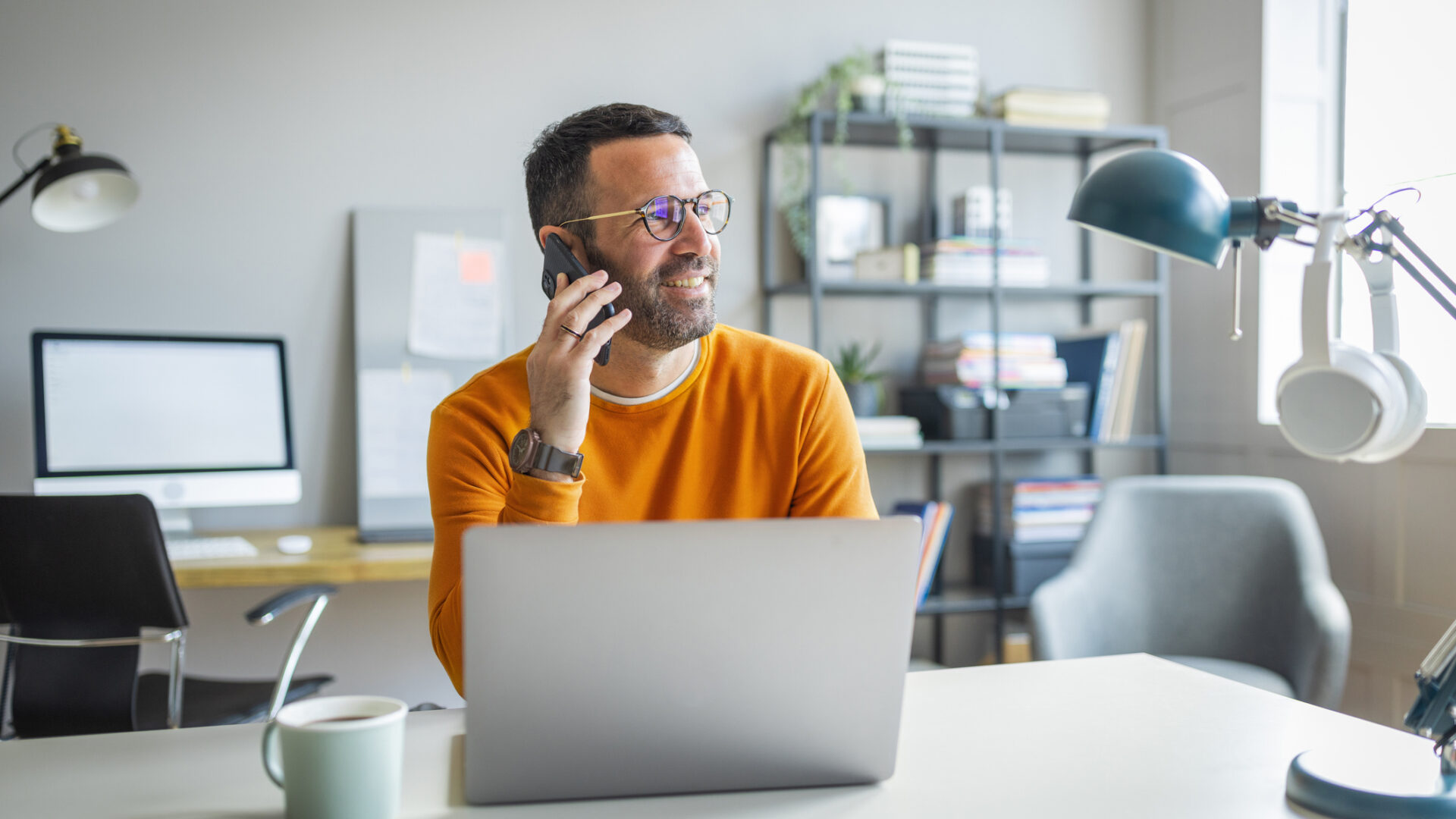 Businessman wearing casual clothing and eyeglasses sitting at his desk, using laptop and smart phone in the office.